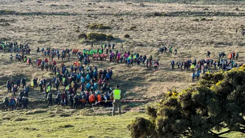 Masses of young people walking on moorland on Dartmoor. There are military personnel wearing high-vis clothing among them.