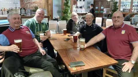Paddy Hennessy Five men sat around a wooden square table smiling and holding pint glasses of beer or larger.