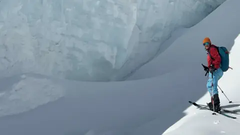 Keith Geddes A skier in bright outdoor gear stands on a snowy slope beside a towering wall of ice, with deep blue sky overhead. The scene highlights the scale of the glacier and the stark contrast between smooth snow and layered ice.