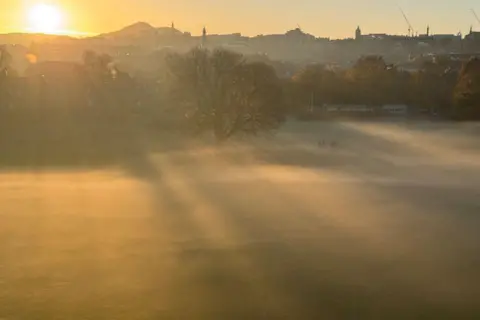 John Edward A misty morning scene at Warriston Recreation Ground, Edinburgh, with the shadows of trees from the sunshine in mist over the ground, as well as shapes of people. The Edinburgh skyline is in the distance.