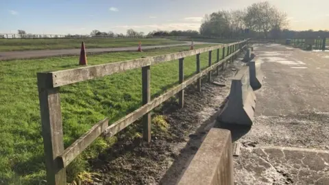 A wooden fence between a racecourse on the left and a road on the right. The nearest panel of the fence is broken.
