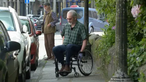 Bob Stride wearing dark trousers and a dark checked shirt, sitting in his wheelchair on a pavement, facing a line of parked cars