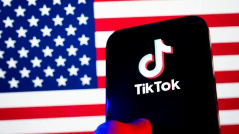 A thumb on a mobile phone using TikTok app with a US flag in the background