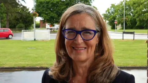 John Fairhall/BBC A woman stands outdoors in the foreground, facing the camera, wearing distinctive blue-framed glasses and a dark top. The background shows a grassy area bordered by a road, with a red car parked to the left and trees filling the scene. A small white sign or structure and a “no entry” road sign are visible further back near the street. The lighting appears natural and even, suggesting an overcast or lightly cloudy day.