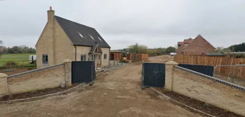 A building site with houses under construction. The entrance of the development with a surrounding wall is in the foreground. The road has not yet had tarmac laid. Construction materials are in the background.