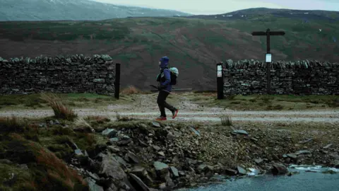 A runner wearing a navy waterproof jacket, black trousers and orange trainers is travelling along the Pennine Way. There is a dry stone wall behind them and to the left of the runner is a stream of water. 