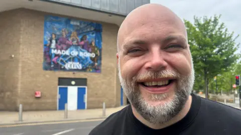 Stephen Irving smiling for the camera with the mural in the background behind him. He is bold and has a short salt and pepper beard. He has blue eyes and is wearing a black t-shirt.