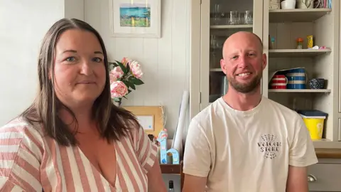 Sarah Doolin wears a pink and white striped top, she had brown hair. Next to her is Craig Pollard wearing a white tshirt. He is bald and has stubble. They are sat next to each other inside a house.