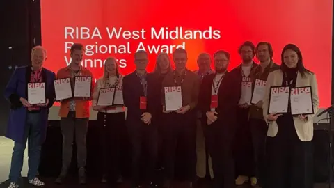 Worcestershire County Council A large group of award winners at the RIBA West Midlands Regional Awards stand together. There are 11 people all stood in formal clothing and some of them are holding up certificates in front of a red background. 