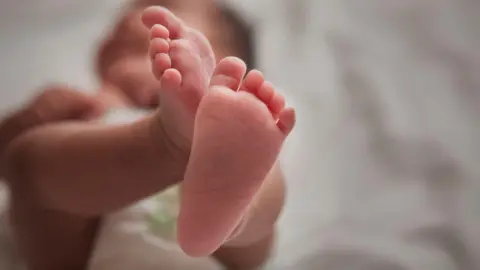 Baby's feet near the camera. There is a blurred image of the baby in the background lying on white sheet.