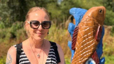 A woman with tied back blonde hair, she is wearing black thick framed sunglasses and a horizontally striped top on. Behind her is a wooden sculpture of a fish.