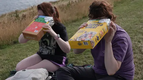 PA Media Two young people are looking into modified cereal boxes to watch the solar eclipse. Sunlight is filtered through a pinprick into the dark box, so the path of the moon across the sun can be seen. One box is Tesco branded Corn Flakes and the other is Shredded Wheat.