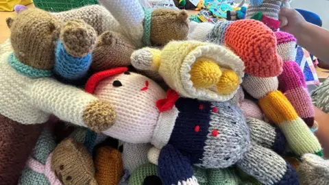 A pile of knitted figures and teddies in brightly coloured wool. A person's hand can just be seen holding one of them up.
