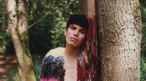 Island Records Jacob Alon leans against a tree in a lush green woodscape. They are wearing a multi-coloured jacket with tassles