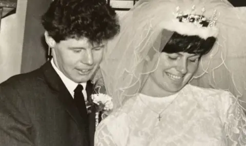 HC-One handout Donald and Margaret Tyman on their wedding day. Donald has short, black curly hair wearing a black wedding suit while Margaret has short black hair wearing a tiara and veil and white wedding dress and gold cross necklace. Both are smiling.