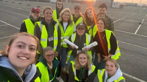 The picture shows a group of people gathered in an outdoor car park area at sunset. They are wearing bright yellow high‑visibility vests, and some have lanyards or passes around their necks. They are standing close together as if posing for a group photo. 