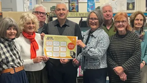 FoCCaL A group of about eight older men and women standing in a library smiling to camera and three of them are holding onto a poster which says 'Book Bingo'