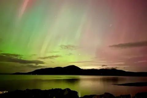 TealKat/BBC Weather Watchers A green and pink aurora over hills and water.