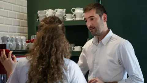 Young woman on her first day at work learning how to use the coffee maker in a coffee shop. She has long curly hair, and her manager, wearing a white shirt looks on.