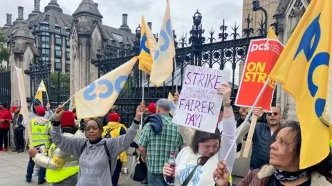 BBC Female PCS union members wave banners saying "Fairer Pay" and "PCS on strike" outside the gates of the Houses of Parliament