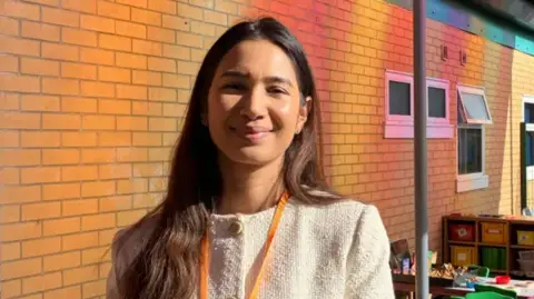 Councillor Adorabelle Amaral Shaikh, who has long dark brown hair and is wearing a cream cardigan, is standing in front of a brick wall at a school with some school furniture in the background.