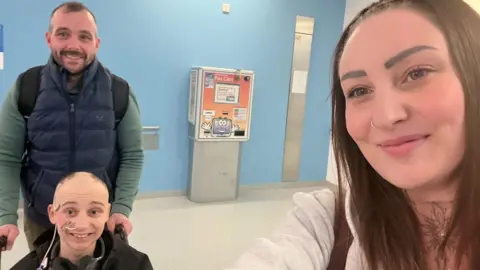 Family photo The boy from the above picture sitting in a wheelchair being pushed by a man wearing a green long-sleeved top under a sleeveless puffer jacket. The boy has lost most of his hair, apart from a few patches. The woman is standing near the camera taking a selfie with them all in.