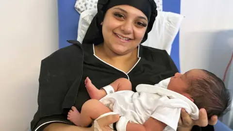 A young woman holds her newborn in her arms and smiles at the camera. 