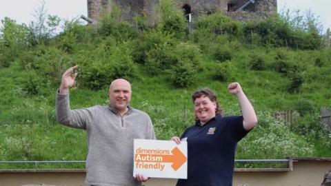 Two people hold an autism friendly sign in front of a castle
