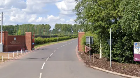 Google The entrance to the Villa training ground, with a road surrounded by lots of green trees and bushes. A red brick wall is on each side of the road with a Villa logo on the wall