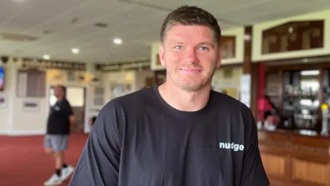 Owen is wearing a black 'Nudge' T-shirt and stands inside a clubhouse with a long wooden bar behind him. The room has red carpeting and display boards mounted above the bar.