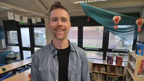 Nick Rees is a middle aged man with grey hair, he has stubble and is wearing a blue shirt. He is standing inside a classroom. 