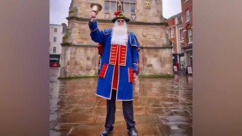 Shrewsbury Town Council A very tall man who has a long white beard is dressed in a town crier outfit. He has a red and blue coat with gold detailing, and black trousers and is ringing a large gold bell. He is wearing a black and gold hat with a red feather.