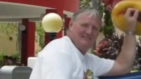 A slightly blurry picture of a man in his 60s with grey hair, wearing a white t shirt.