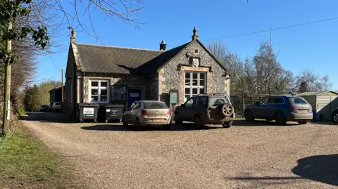 Clare Worden/BBC Great Dunham Primary School is a flint clad building with cars parked in front of it. It is a sunny day, with a completely blue sky above.