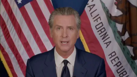 California Governor Gavin Newsom stands in front of the US and the Californian flags.