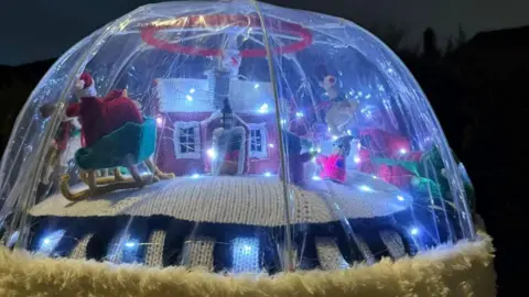 Paula Campbell A scene protected by a clear plastic dome includes a knitted house with pink walls and a snowy, white roof, lit up for Christmas. Santa can be seen to one side, on his sleigh delivering presents. The whole display is lit up with white fairy lights.