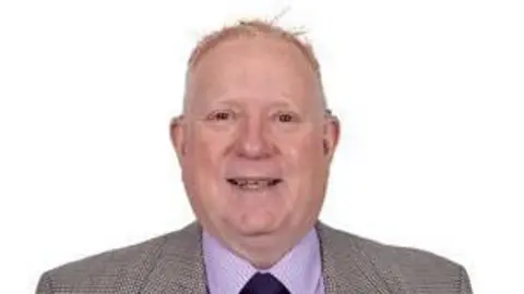 Jamie Hanlon. He is stood in front of a hite background wearing a grey suit and a pink shirt.