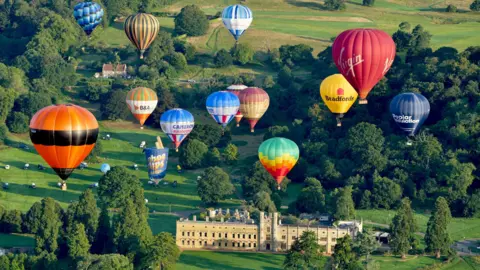 PA Media Several hot air balloons flying over Ashton Court Estate in Bristol. There is a wide, yellow coloured building in the grounds, with many trees around it. The hot air balloons vary in size, shape and colour and they are flying at different heights.

