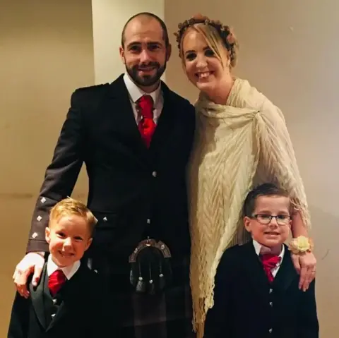 Shelley Martin Barry an Shelley Martin with their twin boys Oliver and Daniel who look to be aged about five in the photo. They are all dressed up as if going to a wedding. Barry and his sons are wearing kilts and formal Scottish attire and Shelley is wearinga cream shawl and flowers in her hair