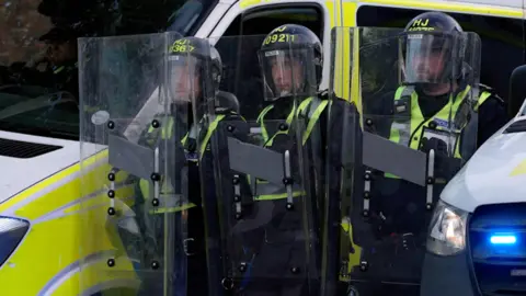 Three police officers in riot gear. They are stood in between two police vans.
