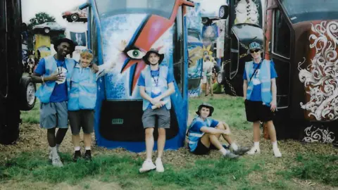 WaterAid A film photo showing five WaterAid volunteers wearing hi-vis blue branded vests and posing for a photo. They are sitting and standing around upturned cars which have been painted, one with a David Bowie style lightening bolt.