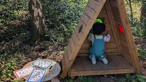 Birmingham Settlement A young girl inside a wooden triangular play den. It is outdoors in a forest. A wooden table with cards is next to the den