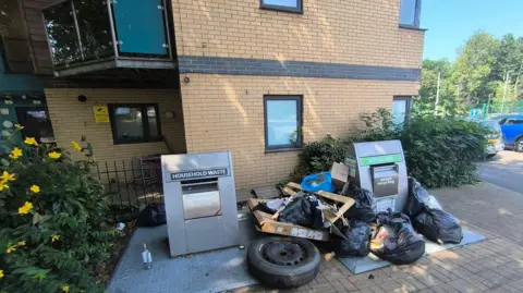 Nuno Bastos Two large silver metal rubbish chutes, with black bin bags placed around them. Behind them is a block of flats, there are some small hedges either side of the bins.