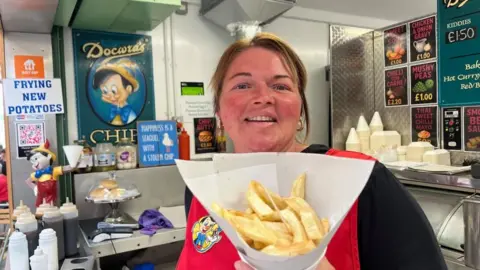 Andrew Turner/BBC Christina Docwra has auburn hair, tied back, and is smiling, holding a cone of chips, standing inside her stall. She is wearing a black jersey and a red tabard. There are various sauces and vinegars, and signage in the background.
