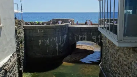 DS Pugh/Geograph Pedestrians walk over a stone bridge crossing the river Lim where it meets the sea. The river is shallow with high stone walls either side with buildings on top. It's a sunny day and the sea beyond the bridge is calm and bright blue.