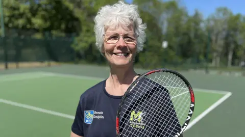 Hilda Moore standing on court smiling wearing a Marlborough Tennis t-shirt holding her racket up to her shoulder