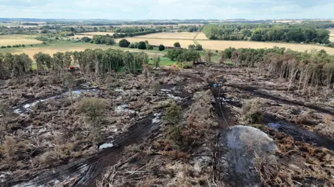 PA Media Birds-eye view of the damaged and burnt nature reserve. Some clouds of white smoke can be seen in the air. There is water being sprayed on to the ground, and in the background a train can be seen passing.