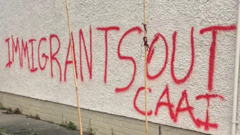 Shows a gable wall with the words "Immigrants Out CAAI" written in red paint