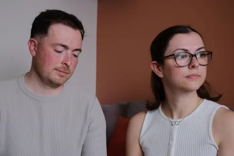 The McCreadys are sitting together on a sofa. Lauryn is looking wistfully out of a window while Andrew is looking downards towards their daughter's memory box