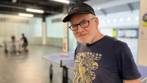 Tom Jackson/BBC Head and shoulders image of Barney McCullagh. He is wearing a black cap and navy t-shirt with a yellow pattern on and is stood in a ping pong venue.